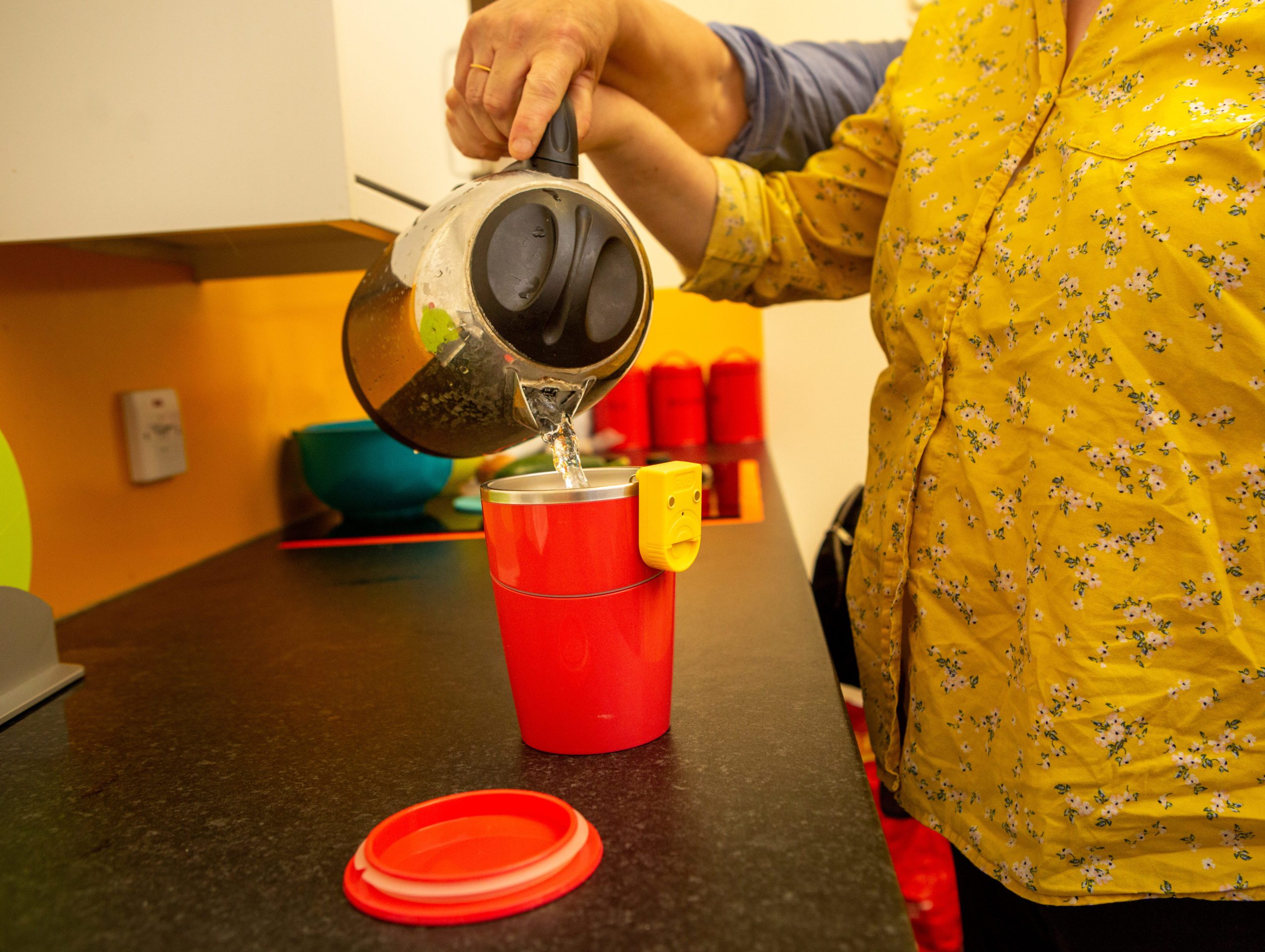 The image shows a persons hands with a kettle of boiling water, they are learning to make a cup of tea using a suction cup and a liquid level indicator.