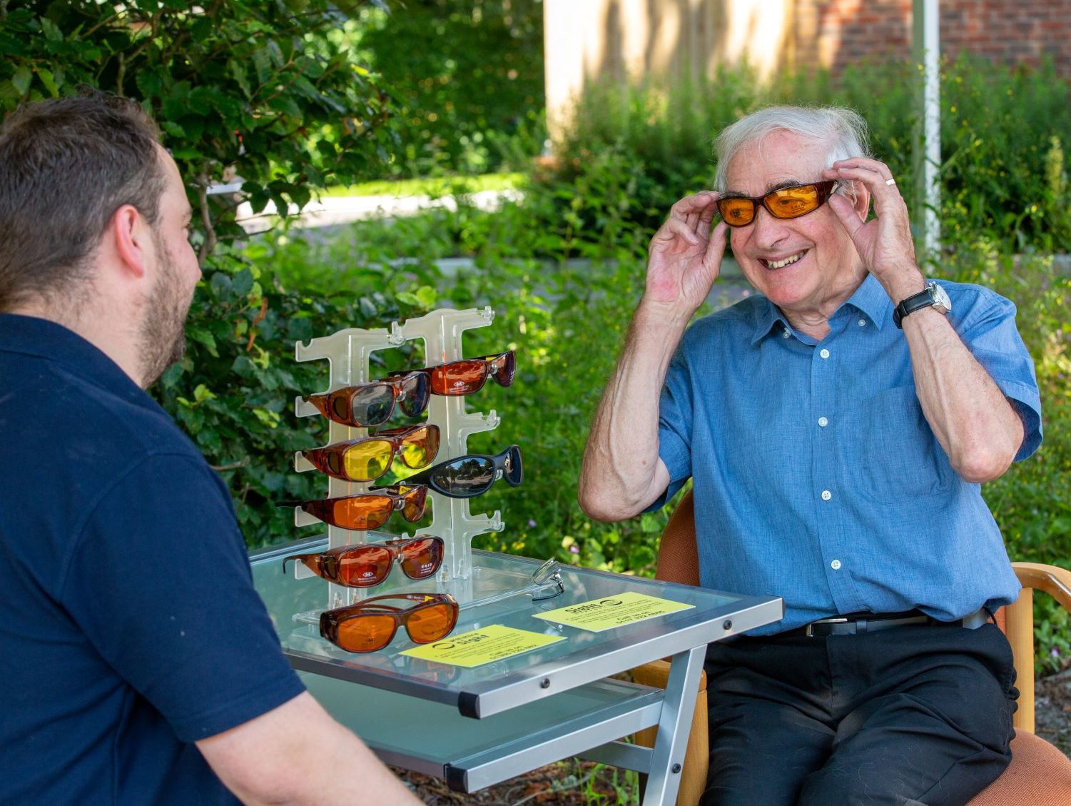 A Community Sight Loss Advisor is sitting outside with a gentleman who is trying out some eyeshields.
