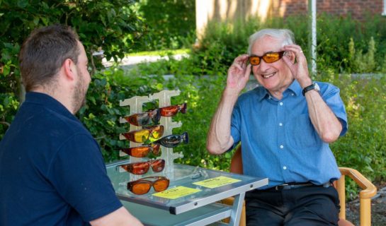 A Community Sight Loss Advisor is sitting outside with a gentleman who is trying out some eyeshields.