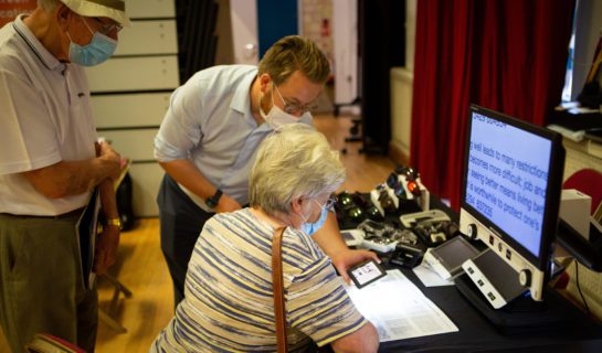 The image shows a lady sat at a table in front of some Hi Tech magnification equipment. A demonstration is taking place.
