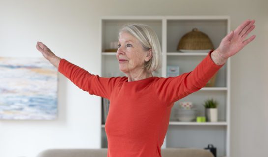 Senior woman exercising in her living room. She has her arms outstretched.