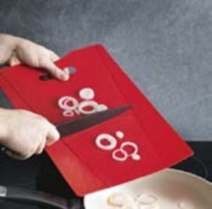 Person pushing white onions into frying pan with knife. The onions are on a red chopping board with raised sides.