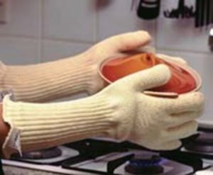 Person holding cake in tin over hob. Person is wearing off white long arm oven gloves.