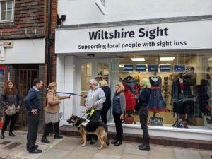 View of the front of Wiltshire Sight Salisbury Charity Shop, outside are 7 people and a guide dog, 3 mannequins are in the window wearing black and red clothes