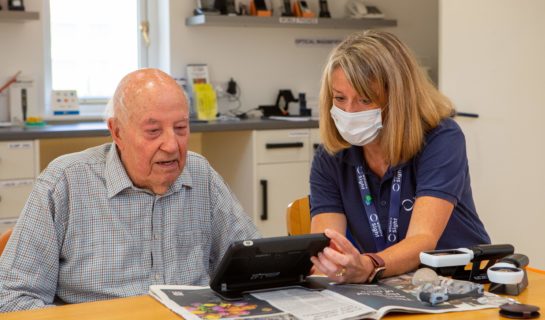 A Sight Loss Advisor is demonstrating an electronic magnifier to a man, they are sat at a desk in our resource centre.