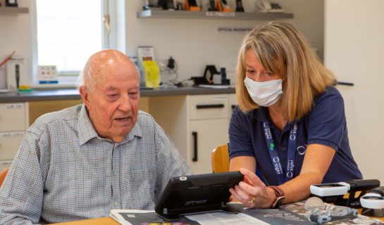 A Sight Loss Advisor is demonstrating an electronic magnifier to a man, they are sat at a desk in our resource centre.