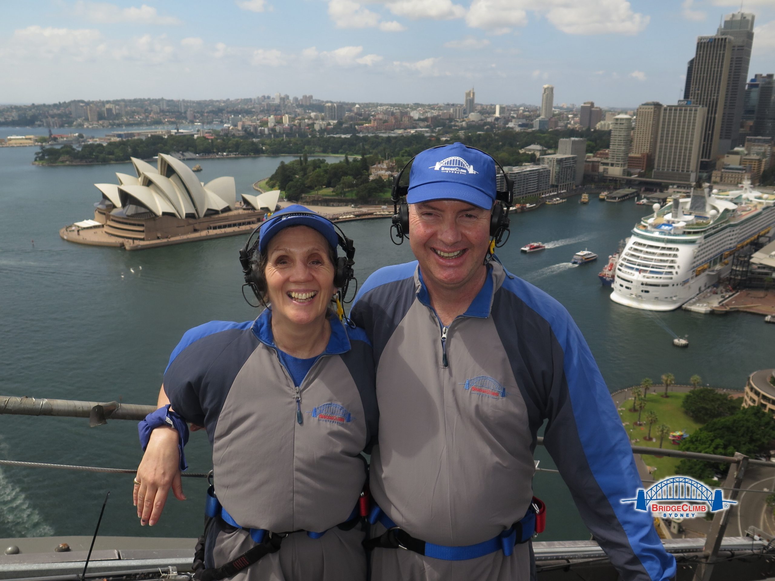 A couple stand at the top of Sydney Harbour Bridge with the Opera House behind them