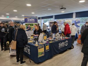 Photo of our stand at Eye Can Bristol. There is a large table with lots of equipmkent and infomation, Staff are talking to visitors.