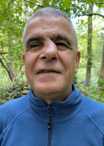 Photo of Kayvan wearing a blue top smiling at the camera, behind him are green trees.