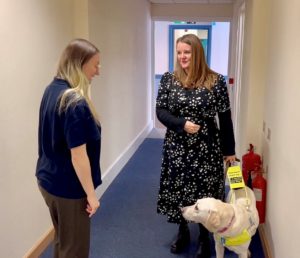 Photo of two women in a corridor, one woman is looking at the guide dog that is guiding the other woman. The guide dog is white adn is looking at the 2nd woman.