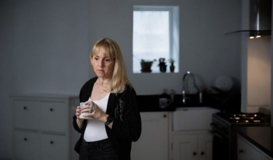 A lady is standing in a darkened room there is a window behind her, she looks sad.