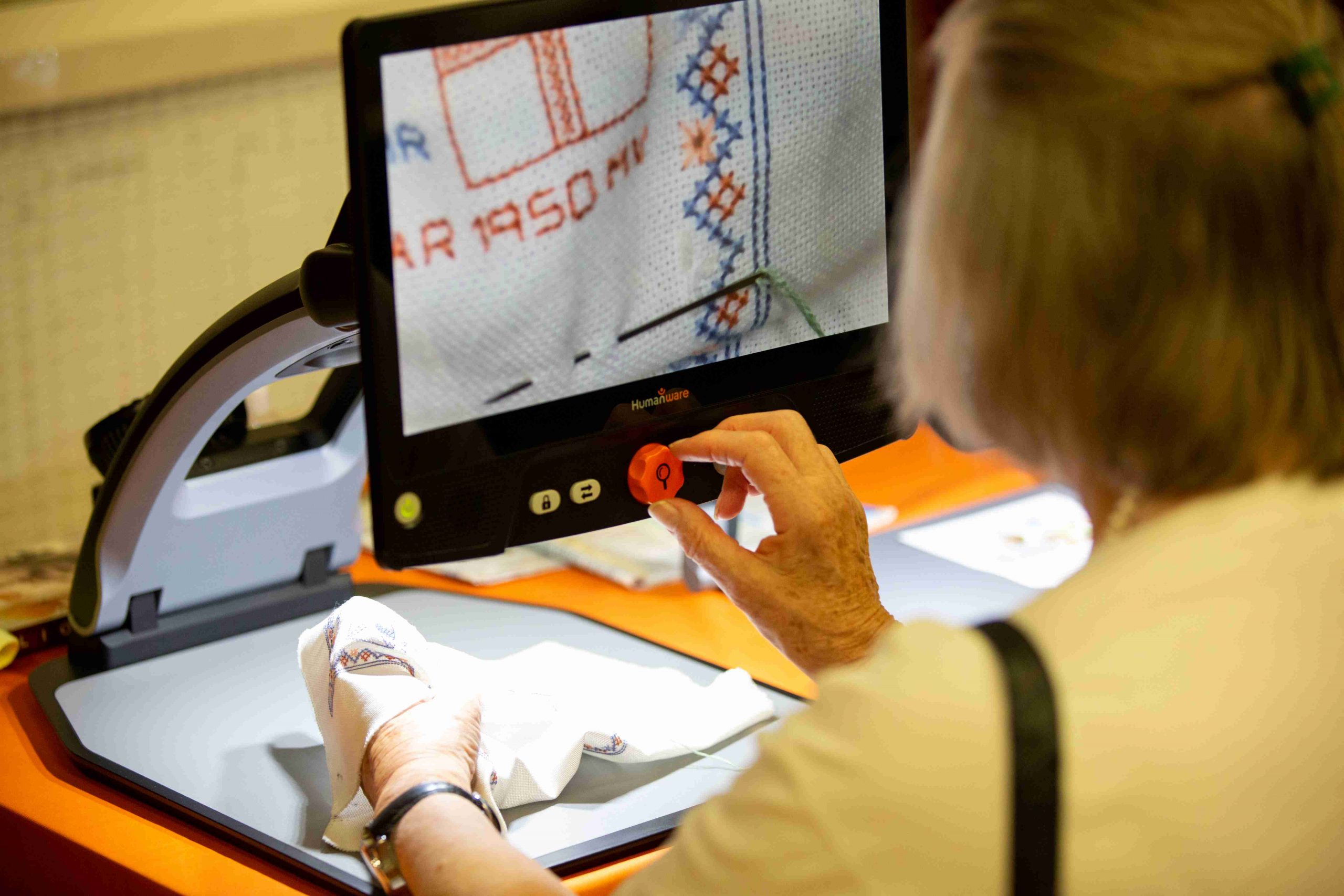 Image shows a woman using an electronic magnifier. She is looking at some embroidery, you can see the needle and thread on the screen in close up.