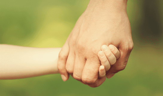 An adult hand holding a child's hand, in the bacground it is green