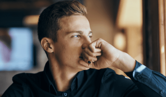 A young white man is looking to the right, he has his hand to his mouth and he is looking thoughtful.