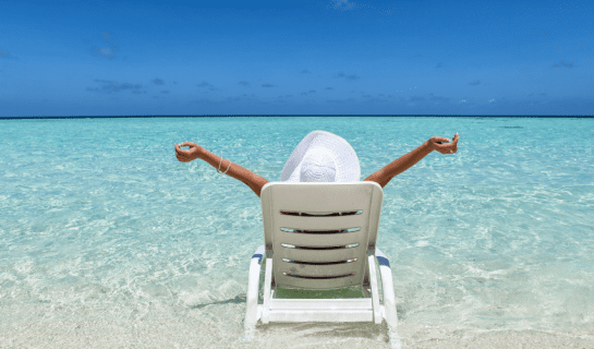 A person is sat on a white deckchair with a white hat on and arms outstretched, they are surrounded by crystal clear water and are facing a blue sky and sea horizon