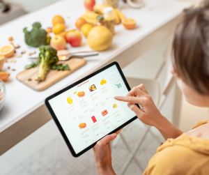 image of someone online shopping on a tablet, they are sitting in front of a table with vegetables on and they are shopping for vegetables on the tablet.