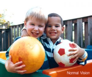 Two boys are smiling at the camera, they are both holding footballs, behind them is a fence. One boy is white with blond hair and the other boy is of colour.