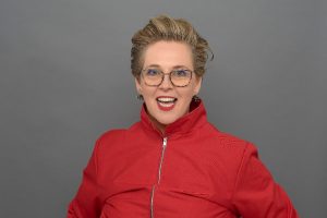 Louise is facing the camera smiling, she is a white lady with blonde short hair, glasses bight red lipstick and is wearing a bright read zip up top. 