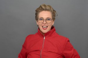 Louise is facing the camera smiling, she is a white lady with blonde short hair, glasses bight red lipstick and is wearing a bright read zip up top.