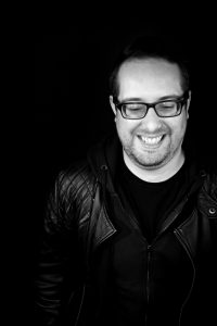A black and white photo. Jake is wearing dark framed glasses and black clothing, he is smiling at the camera. Behind the background is black.