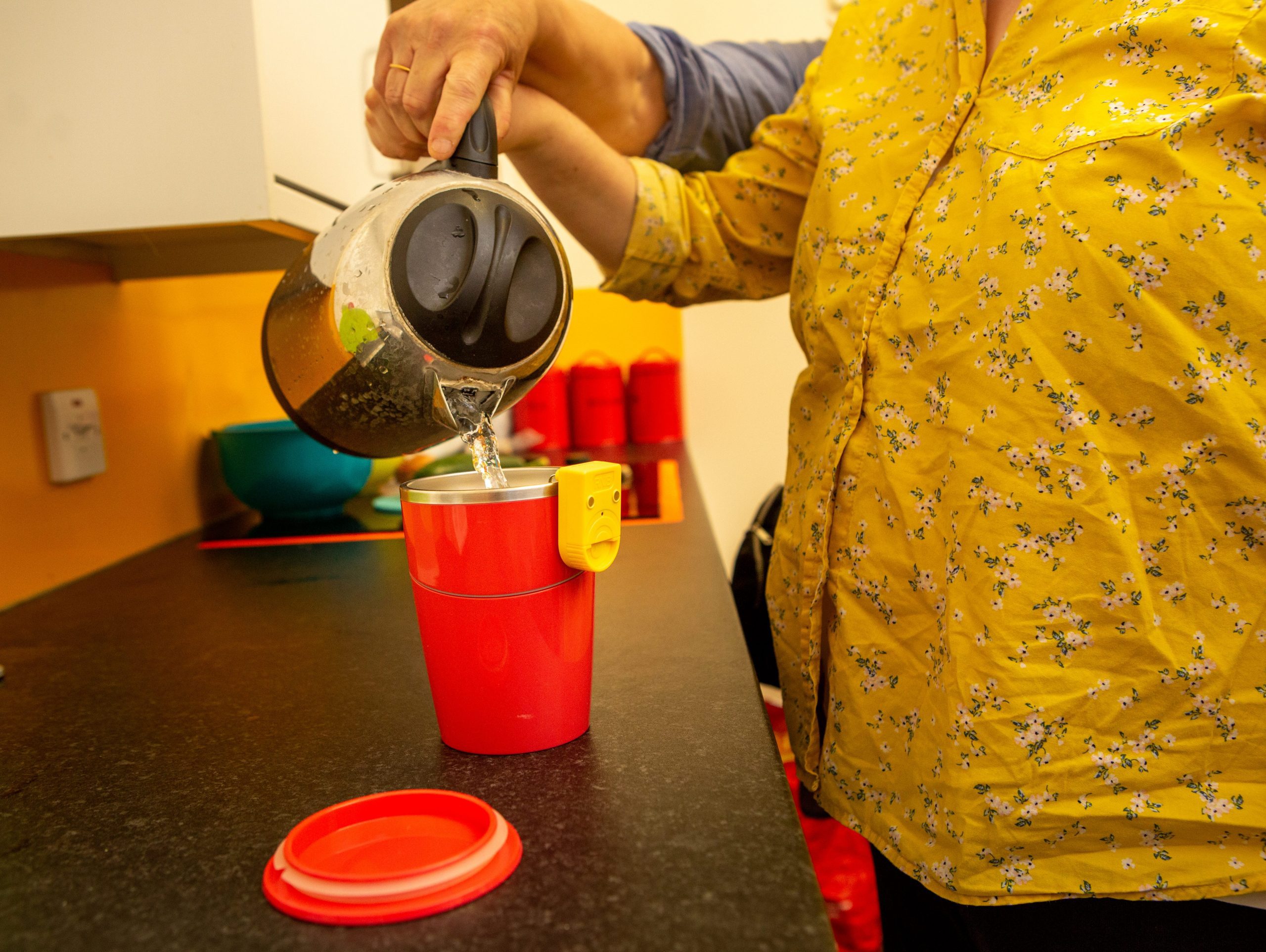 The image shows a persons hands with a kettle of boiling water, they are learning to make a cup of tea using a suction cup and a liquid level indicator.