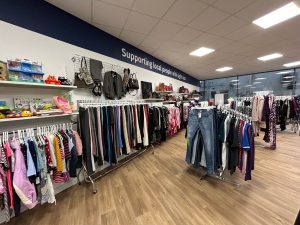 A white wall is full of clothing on shelves and rails. A dark blue banner of paint is at the top. White text reads Supporting local people with sight loss.