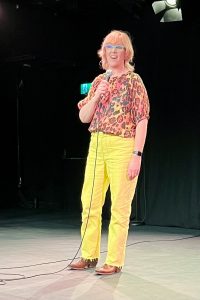 Louise Leigh speaking into a mic. She is wearing yellow trousers and a patterned top.