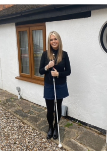 Claire Sisk is wearing black and standing agains a white wall outside, She has her cane Rick held in front of her.