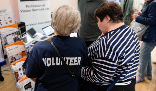 A photo from one of our events. From the back: a lady wearing one of our Volunteer t-shirts is assisting a lady by guiding and explaining the equipment in front of them.