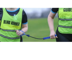 Two men running wearing high vis vests, one says Blind Runner and one says Guide Runner. They are both holding a rope strap to tether together.