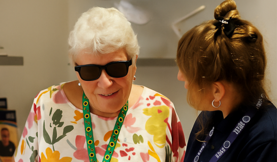 A woman wearing smart glasses and a “Partially Sighted” lanyard smiles while speaking with a Sight Support staff member in a resource centre, with assistive phones and brochures visible in the background.