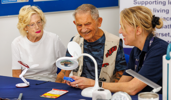 A Sight Loss Advisor demonstrating equipment to an older man and woman at a resource centre table, with magnifiers and lamps on display.