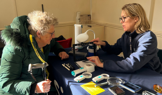 A Sight Loss Advisor demonstrates equipment to a service user at a community hub table, with magnifiers and low vision aids laid out in front of them.