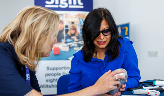 A Sight Loss Advisor showing a piece of assistive equipment to a woman wearing dark glasses at a resource centre.