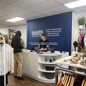 Inside the new Insight Gloucestershire charity shop in Stroud. A staff member stands behind the white till counter serving a customer. The wall behind is painted dark blue with the words: “Insight Gloucestershire – If you or someone you know is experiencing sight loss ask us how can we help.” Clothing and shoes are displayed around the till area.