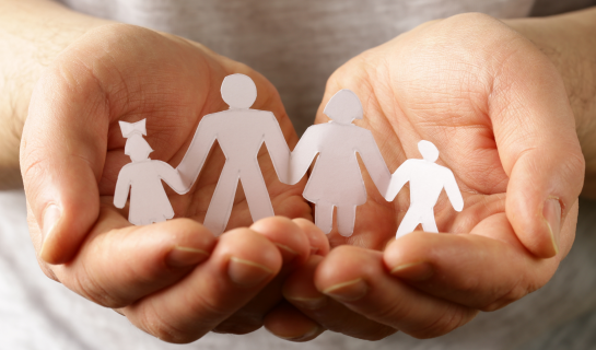 Close-up of a person’s hands gently holding paper cut-outs of a family — two adults and two children