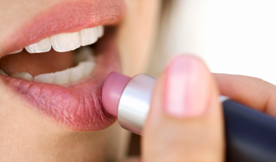 A close-up of a person applying light pink lipstick to their lips. The image focuses on the hand holding the lipstick and the soft natural texture of the lips