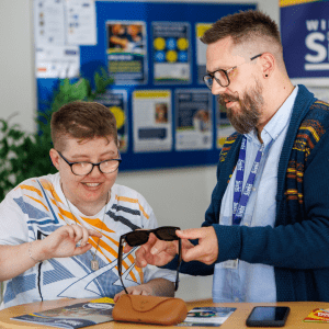 A Digital Skills Advisor demonstrates Meta smart glasses to a smiling person seated at a table, showing how the glasses work during a one to one support session.