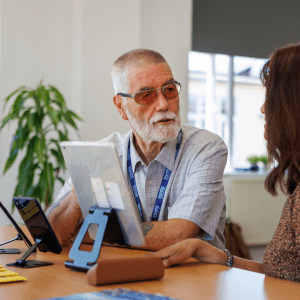 A Techbuster volunteer sits at a table talking through technology options with a person, using a tablet on a stand to demonstrate accessible digital tools during a one to one support session.
