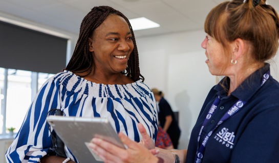 Two women smiling and talking at a hub, with one holding a tablet while a Sight Support staff member explains something, showing friendly one to one digital support.