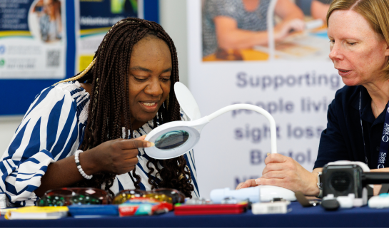An advisor and lady are sitting at a hub table exploring a lighted magnifier. There is other equipment on the table.