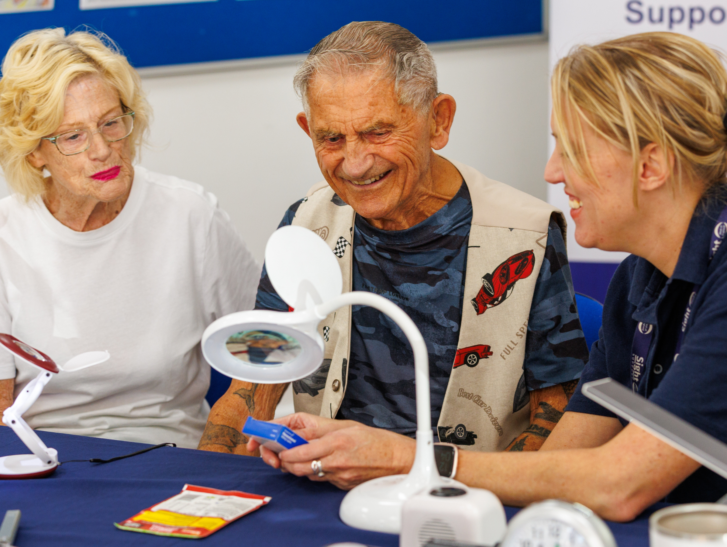 A Sight Loss Advisor demonstrating equipment to an older man and woman at a resource centre table, with magnifiers and lamps on display.