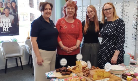 Four women at a bake sale in an opticians. With a table of delicious cakes in front of them.