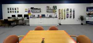 Our Trowbridge accessible Resource Centre with a large table and chairs in the foreground. Shelves and counters around the room display a wide range of sight loss equipment and technology, including electronic magnifiers, lighting, phones and everyday living aids.
