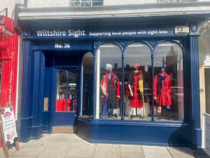 Exterior of the Wiltshire Sight charity shop in Devizes, with a dark blue shopfront and large display window featuring mannequins dressed in colourful clothing. The sign above reads “Wiltshire Sight – Supporting local people with sight loss.”