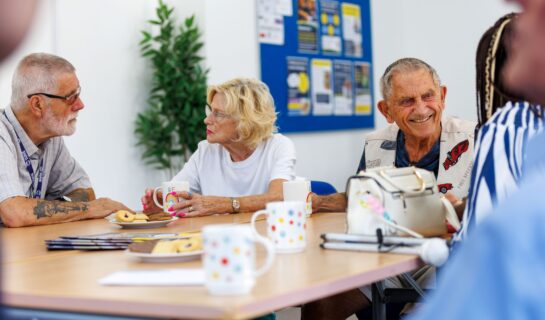 A group of older adults enjoying a relaxed social gathering around a table. A man and woman chat over cups of tea, while another man smiles warmly. Mugs, biscuits and information leaflets are spread across the table, creating a friendly and welcoming atmosphere.