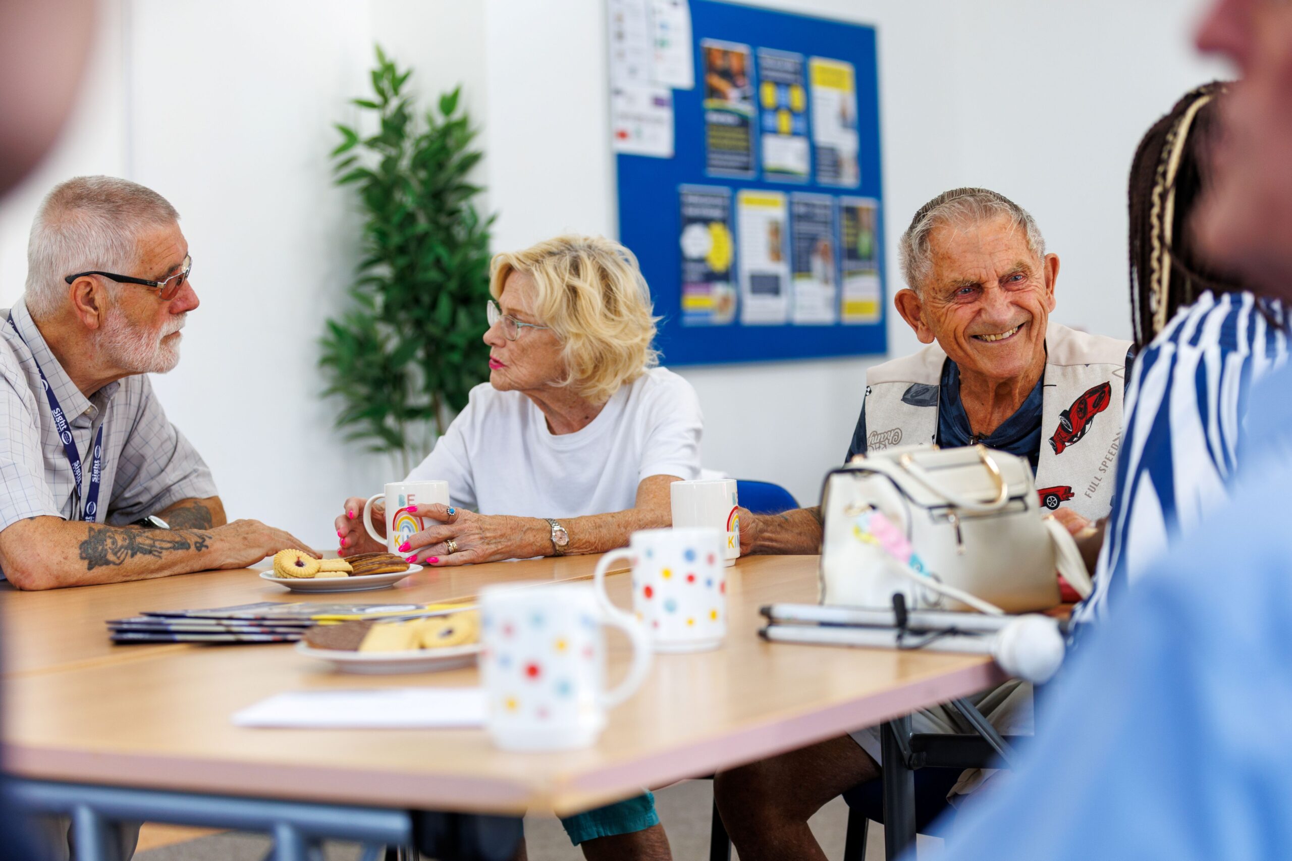A group of older adults enjoying a relaxed social gathering around a table. A man and woman chat over cups of tea, while another man smiles warmly. Mugs, biscuits and information leaflets are spread across the table, creating a friendly and welcoming atmosphere.