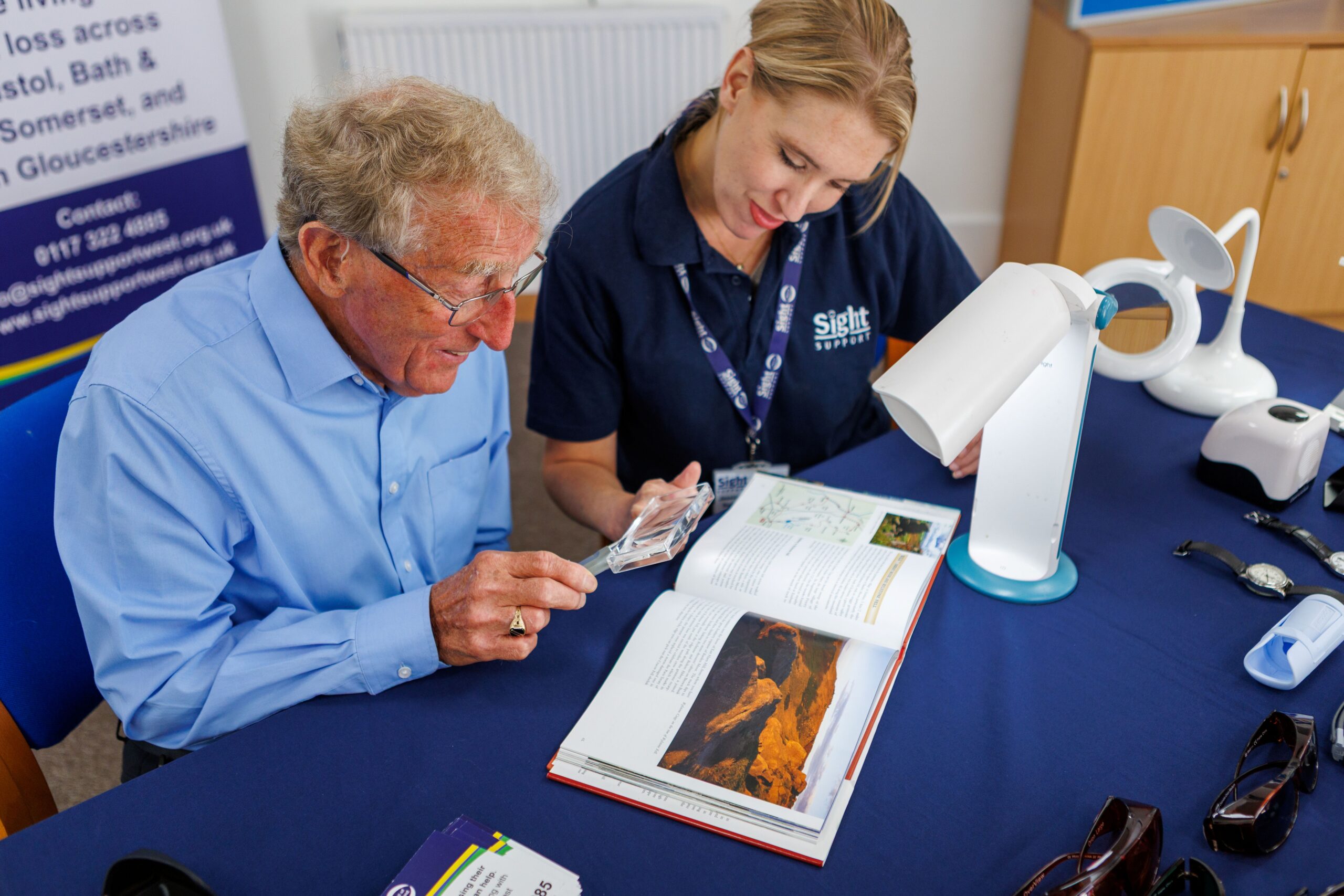 An man wearing glasses uses a handheld magnifier to read a book, while a female Sight Support advisor demonstrates specialist magnification equipment beside him. They are seated at a table displaying a range of visual aids, including illuminated magnifiers, glasses and other equipment designed to help people living with sight loss.