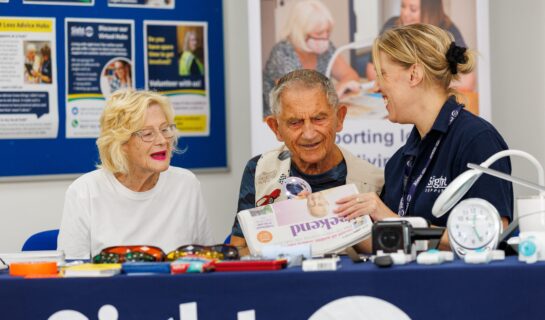 A smiling Sight Support advisor demonstrates a handheld magnifier to a man and woman seated at a table. The man holds a newspaper while the woman looks on with interest. A range of specialist visual aids and everyday living equipment is displayed on the table, including magnifiers, talking clocks and reading aids.