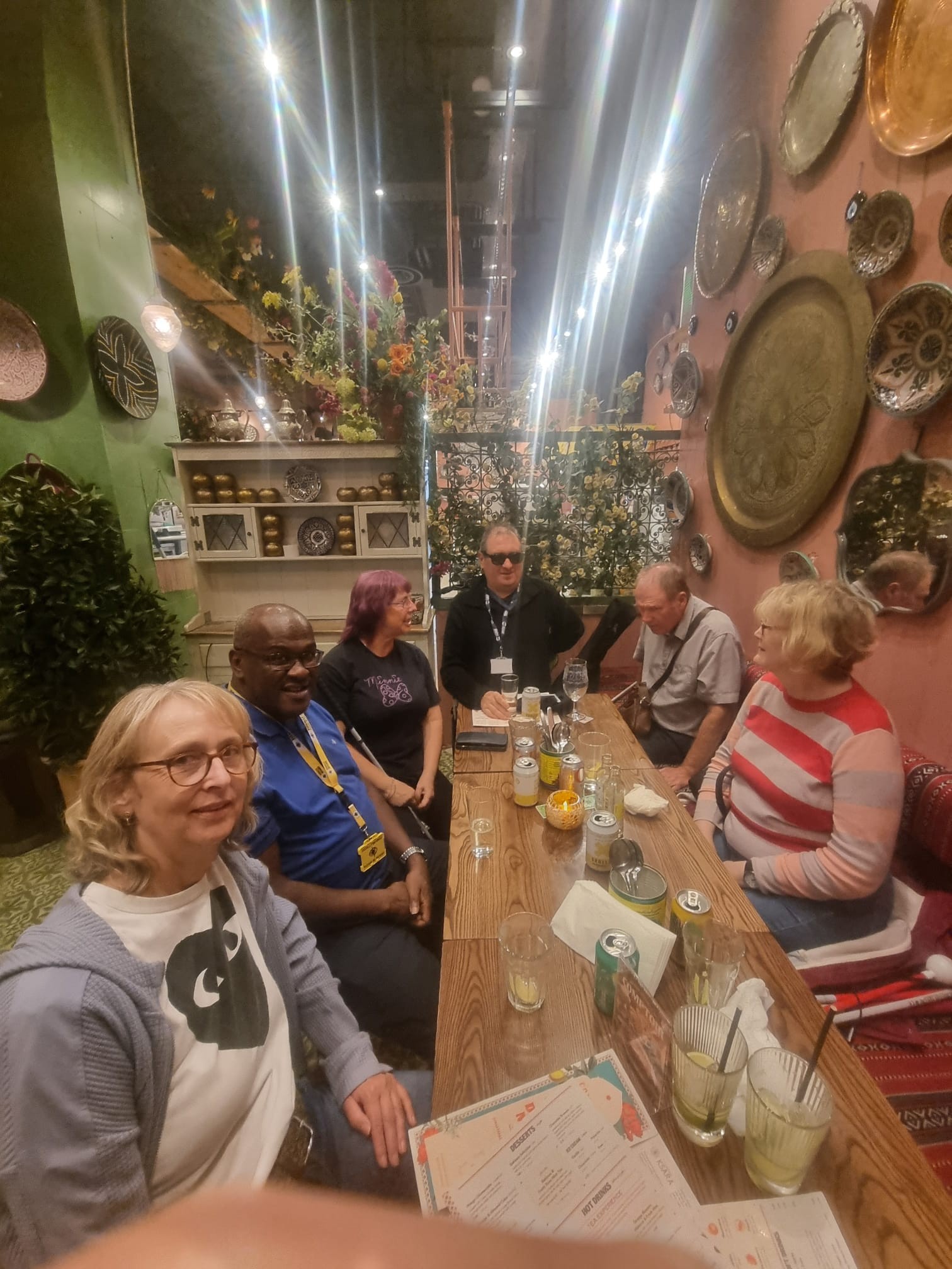 A group of six people sitting together around a long wooden table at a social gathering in a colourful café. Drinks are on the table, and the group are chatting and smiling. The café has warm lighting, floral displays and decorative plates on the walls, creating a welcoming and relaxed atmosphere.